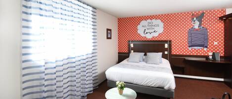 A hotel room with a bed, a small round table with a plant, a grey sofa, and a polka dot wall with a cartoon character.