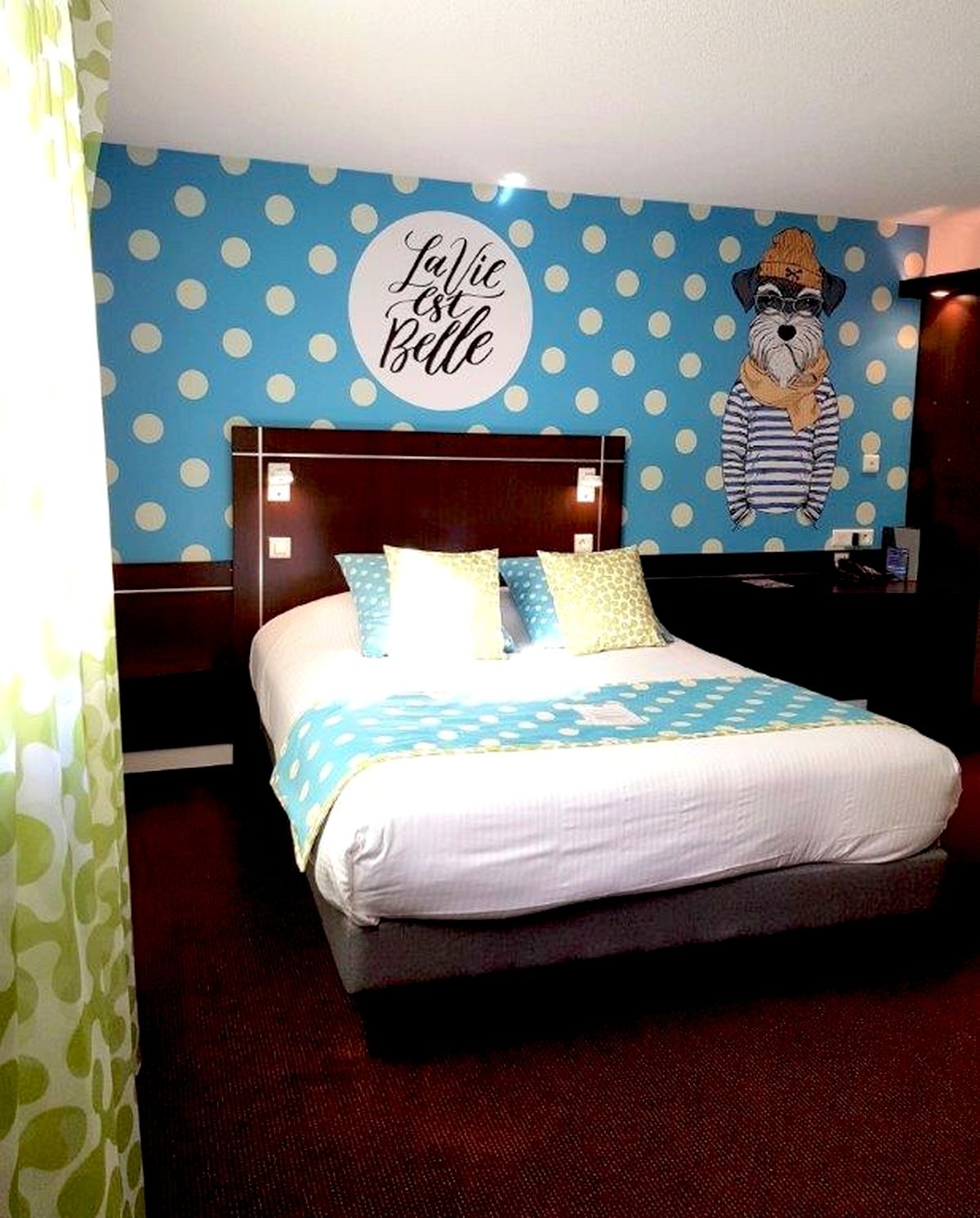 A hotel room with a bed, a headboard, a wall with a polka dot pattern, and a wall decoration featuring a dog.