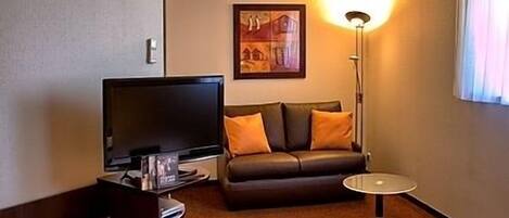 Habitación de hotel con televisión de pantalla plana, sofá de cuero con cojines naranjas, una mesita redonda y una lámpara de pie.