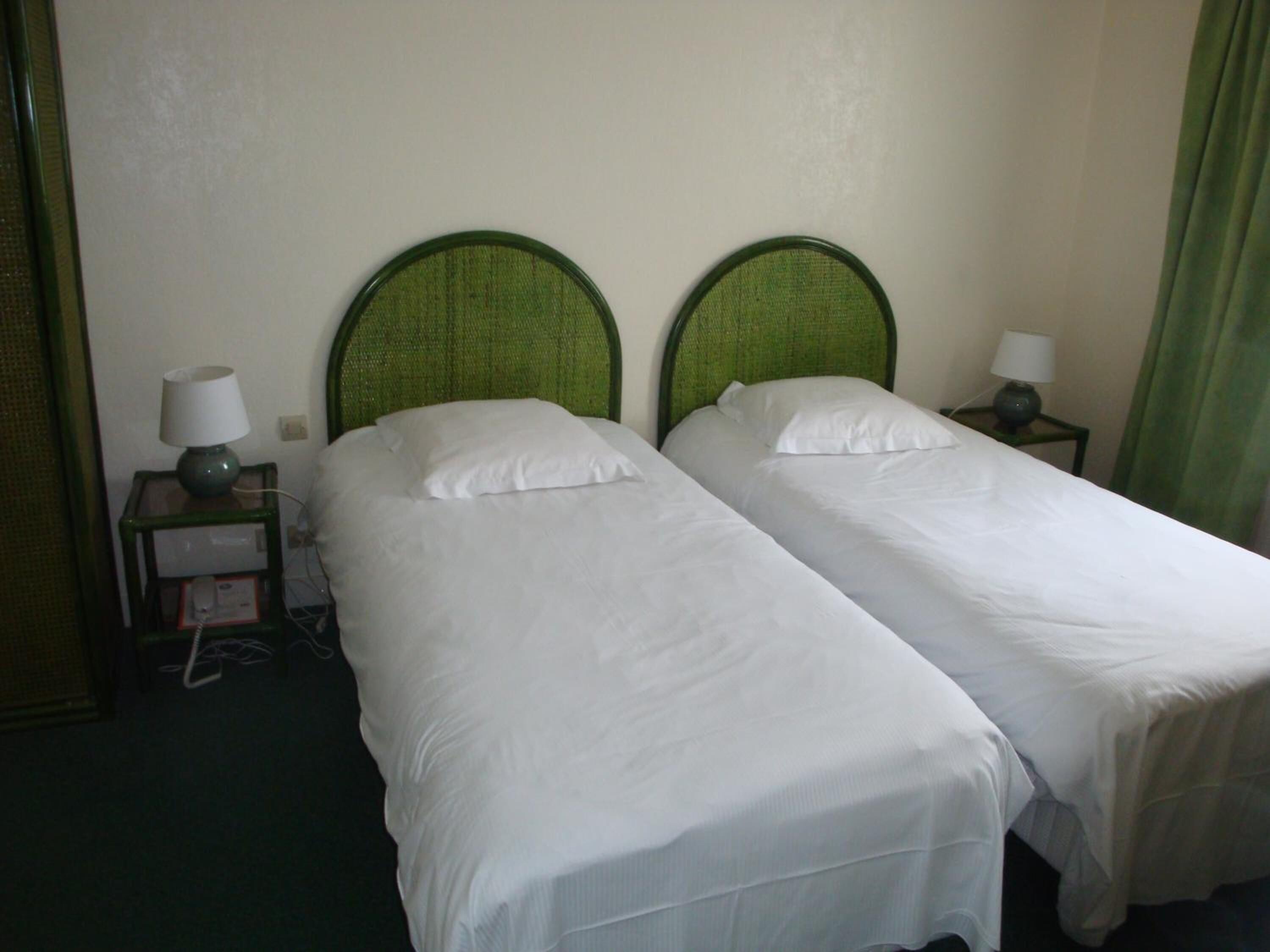 Habitación de hotel con dos camas individuales, cada una con cabecero verde, ropa de cama blanca y una mesita de noche con lámpara.