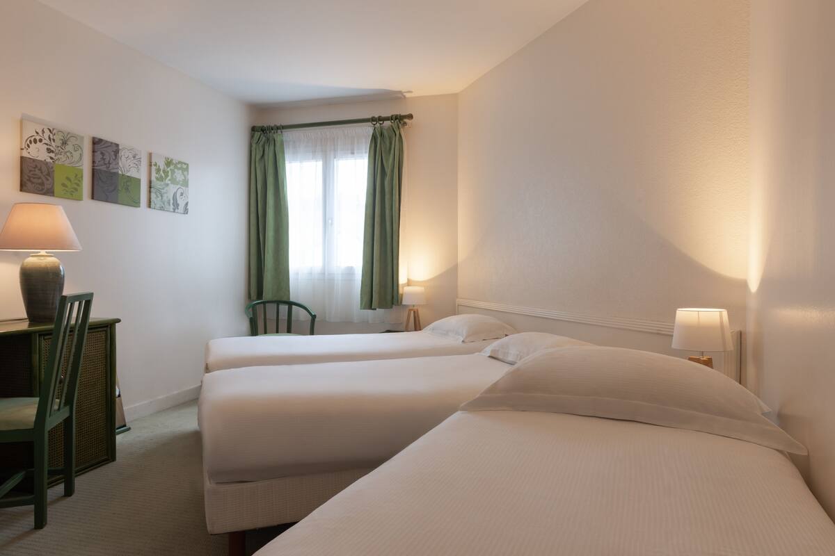 Habitación de hotel con dos camas, un escritorio con lámpara, una silla y una ventana con cortinas verdes.