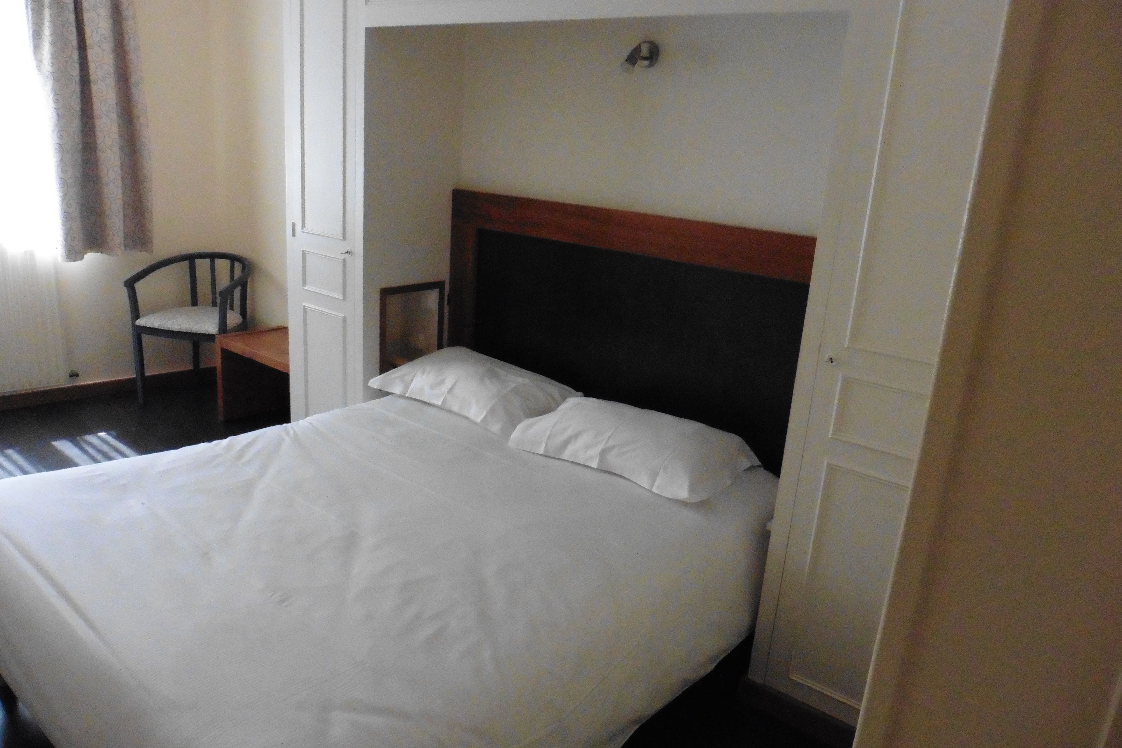 Una cama con sábanas blancas, cabecera de madera y una silla en una habitación con puerta y un interruptor de luz en la pared.