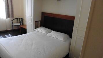 Una cama con sábanas blancas, cabecera de madera y una silla en una habitación con puerta y un interruptor de luz en la pared.