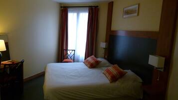 A hotel room with a bed, a chair, a desk, and a window with curtains.