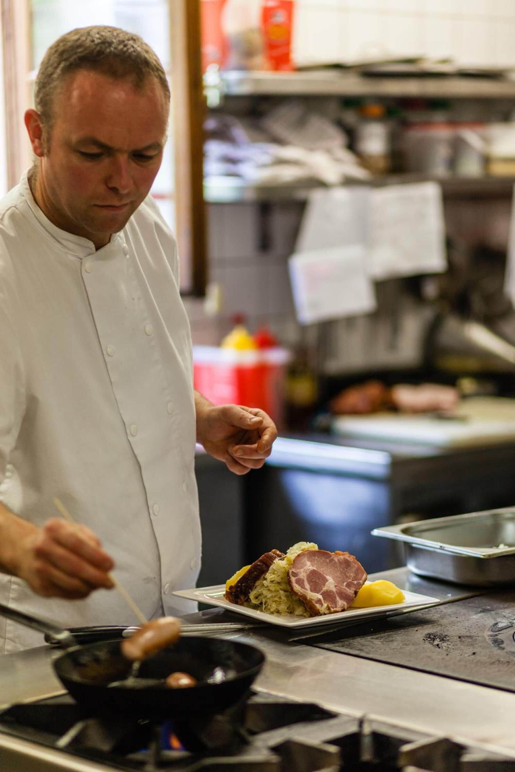 A chef cooking in a professional kitchen with a stove, a frying pan, and a plate of food.