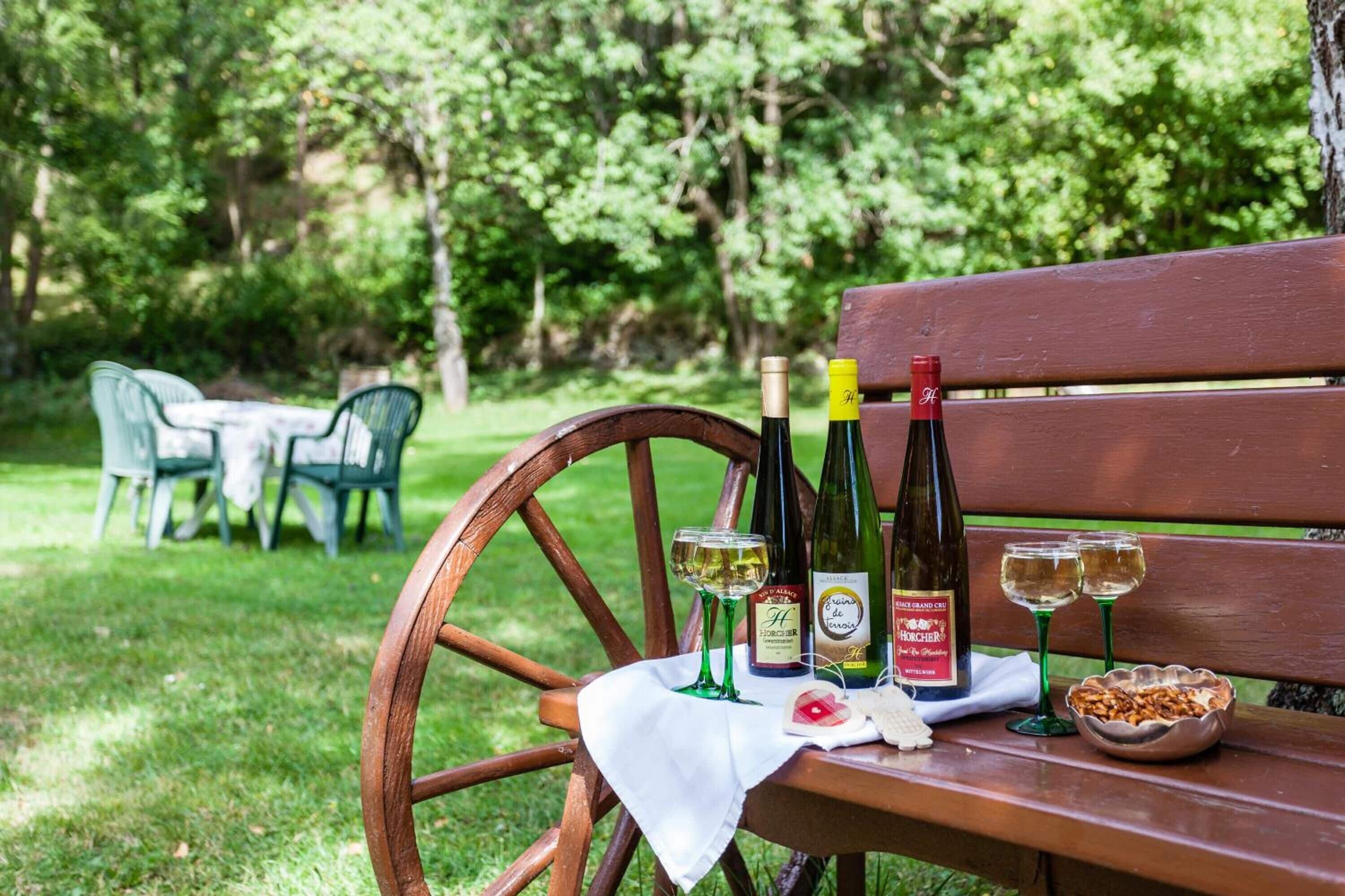A wooden bench with a white cloth, two wine bottles, two wine glasses, and a bowl of nuts in a park setting.