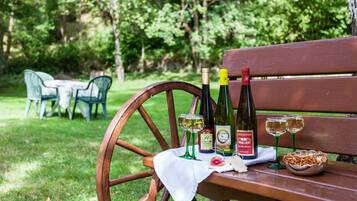 A wooden bench with a white cloth, two wine bottles, two wine glasses, and a bowl of nuts in a park setting.