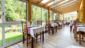 Un restaurante con vigas de madera y grandes ventanales que dan a un jardín. Las mesas están dispuestas con manteles blancos y servilletas a cuadros rojos y blancos.
