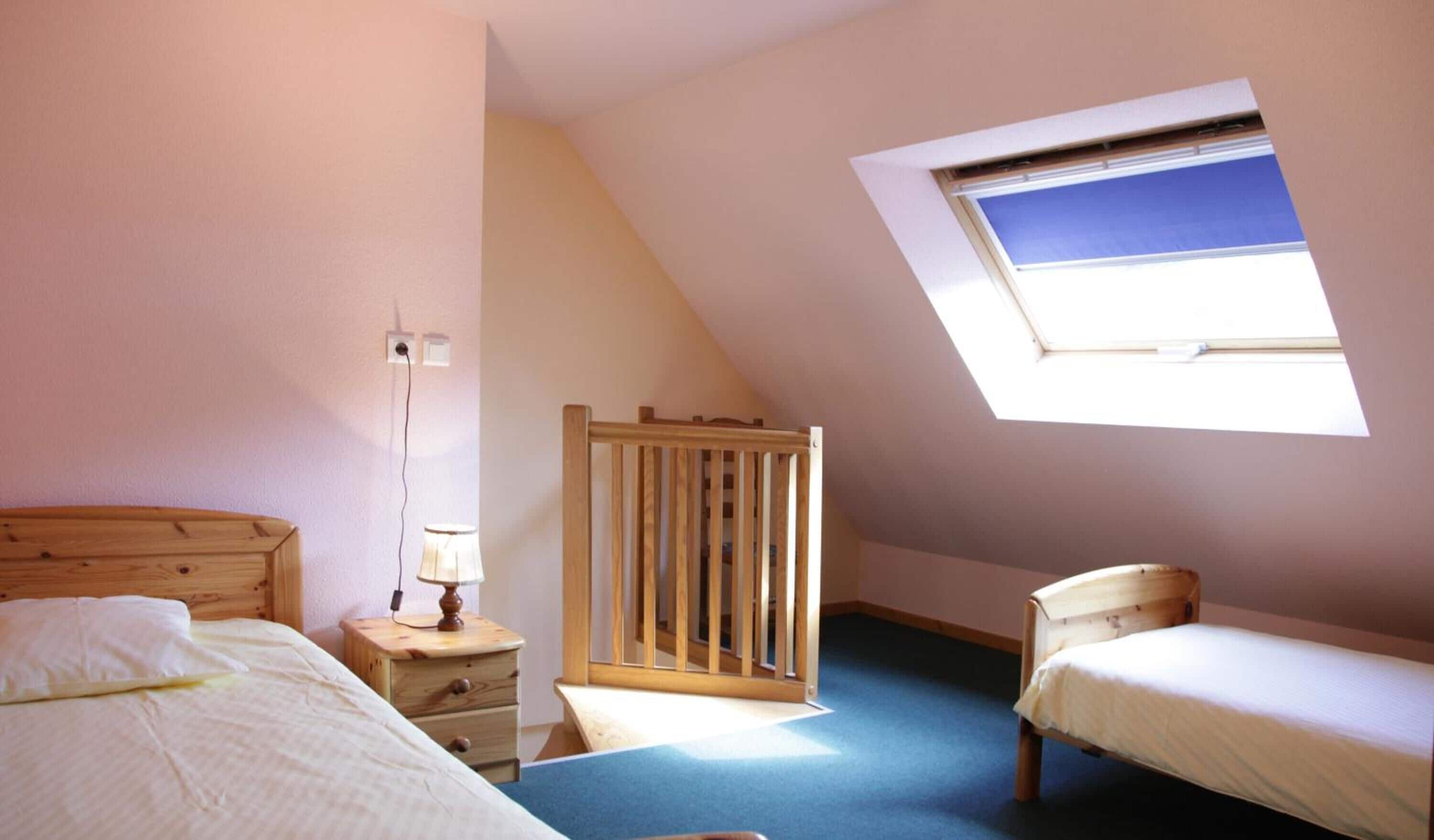 Un dormitorio con techo inclinado, tragaluz, cama de madera, mesita de noche con lámpara y una silla pequeña.