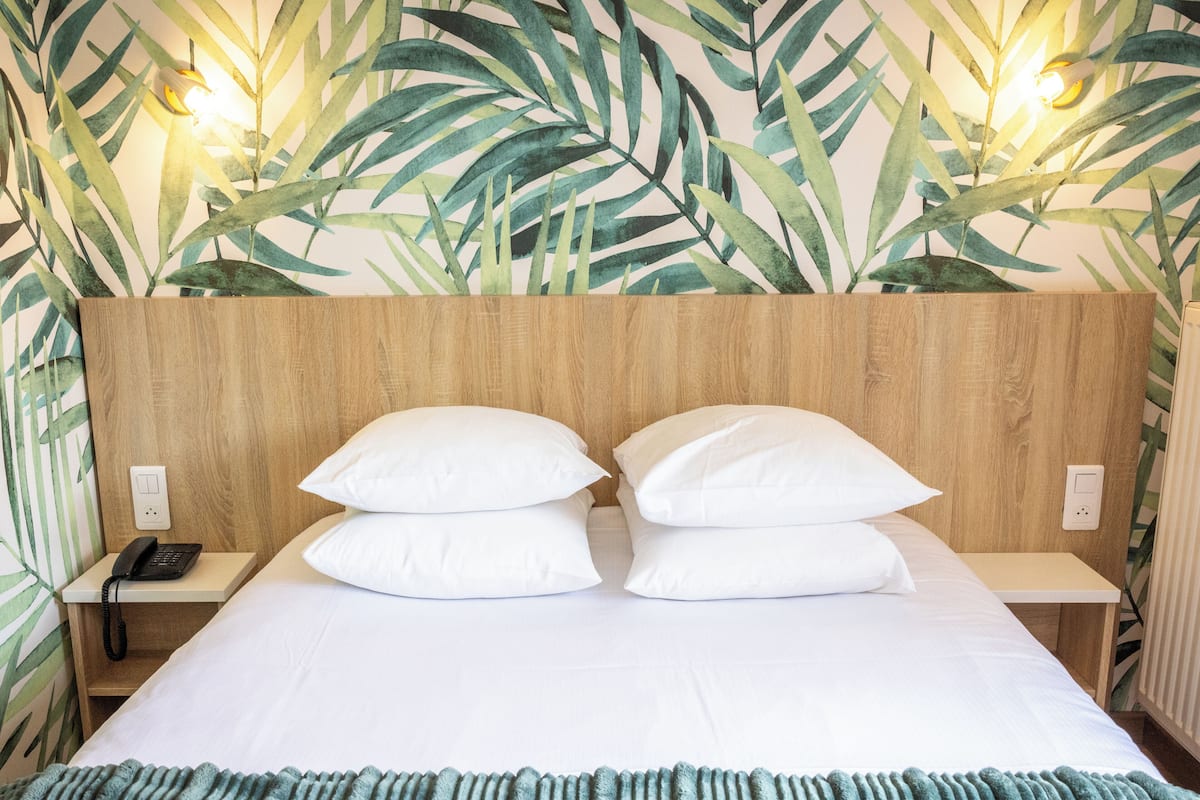 A hotel room with a wooden headboard, two white pillows, a teal blanket, and a wall with a tropical leaf pattern.