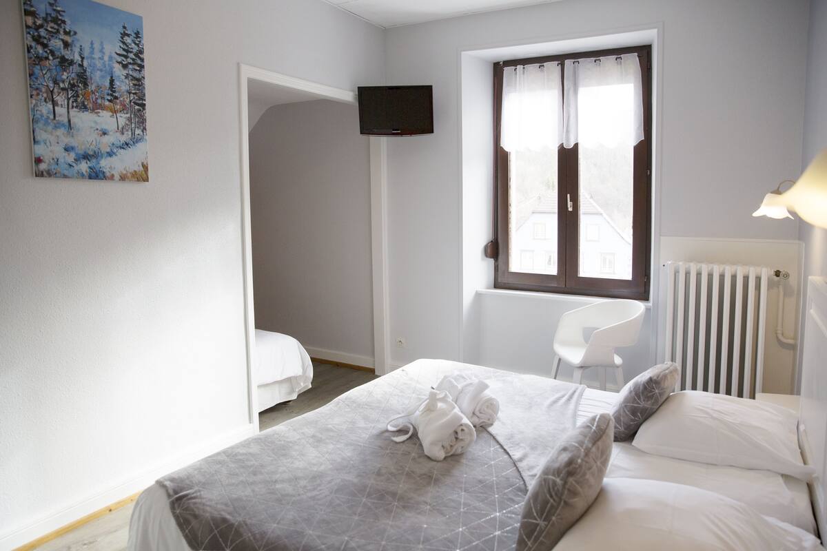 A hotel room with a bed, a chair, a window with curtains, a painting on the wall, and a television mounted on the wall.