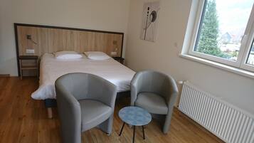 Un dormitorio con una cama, dos sillones, una mesita y una ventana que ofrece vistas a casas.