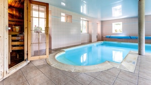 Una piscina interior con estructura de madera en un lateral, paredes con azulejos y agua de color azul cristalino.