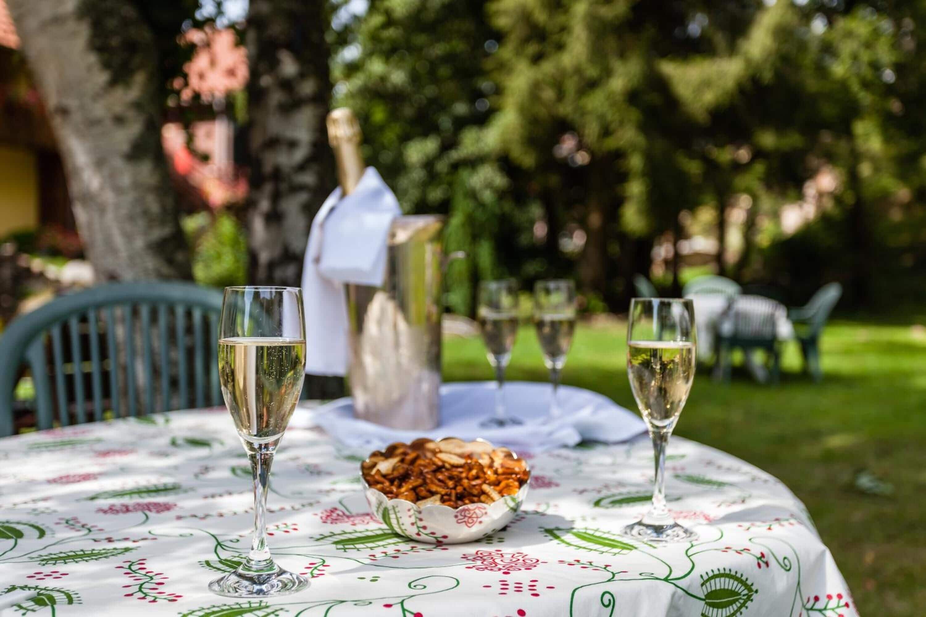 A round table with a floral tablecloth, two wine glasses, a bottle of champagne, and a bowl of nuts.