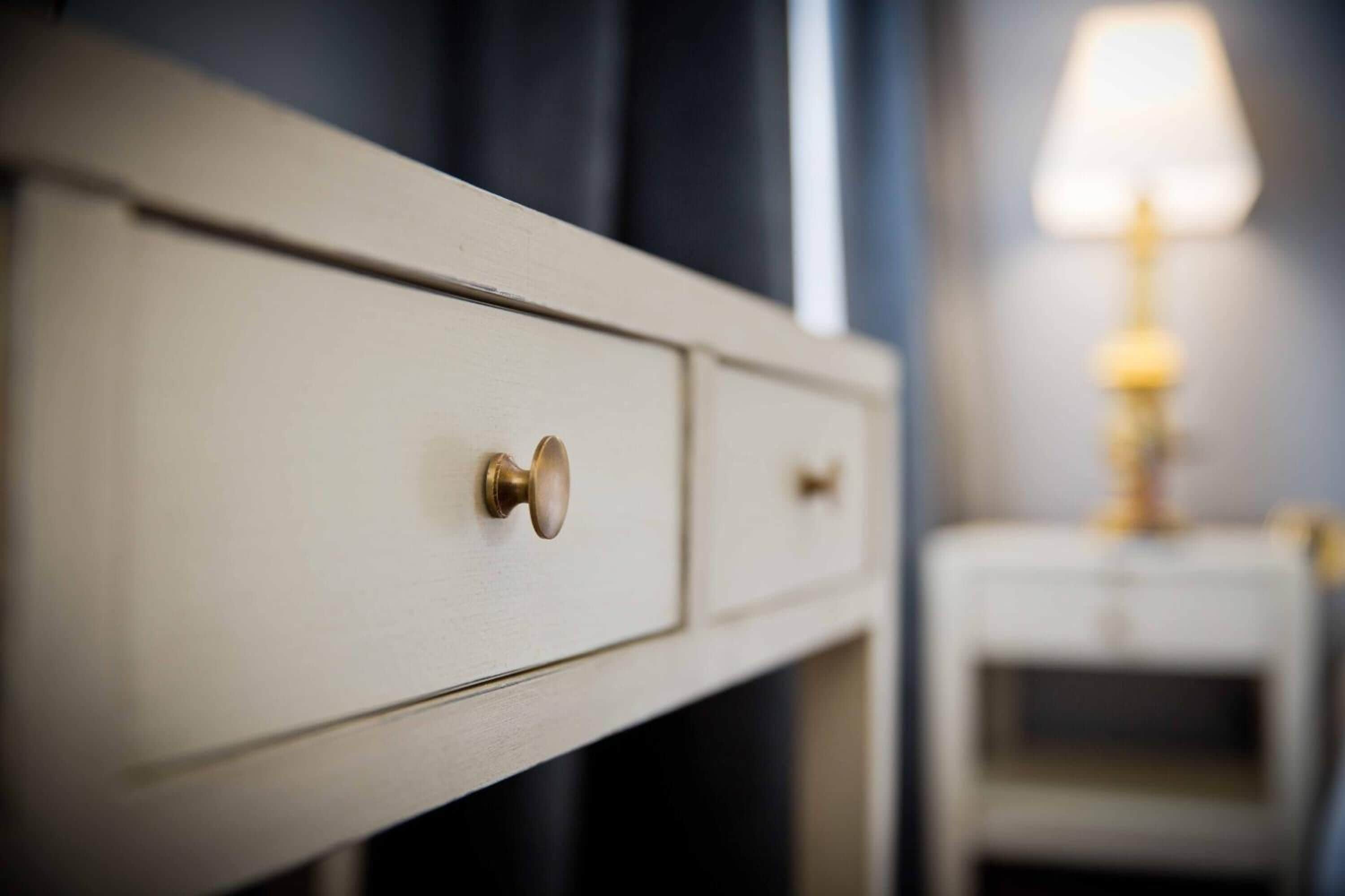Eine Nahaufnahme eines weißen Schminktisches mit einer Schublade mit Messinggriff, eines kleinen Nachttisches mit einer Lampe und einer dunkelblauen Wand im Hintergrund.