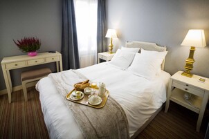 A neatly made bed with white linens, a bedside table with a lamp, a small desk with a chair, and a tray with breakfast items.