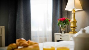A neatly made bed with a white pillow and a white headboard. A bedside table holds a glass of orange juice, a glass of water, and a small vase with red flowers. - Hôtel Champerret Héliopolis (Paris)