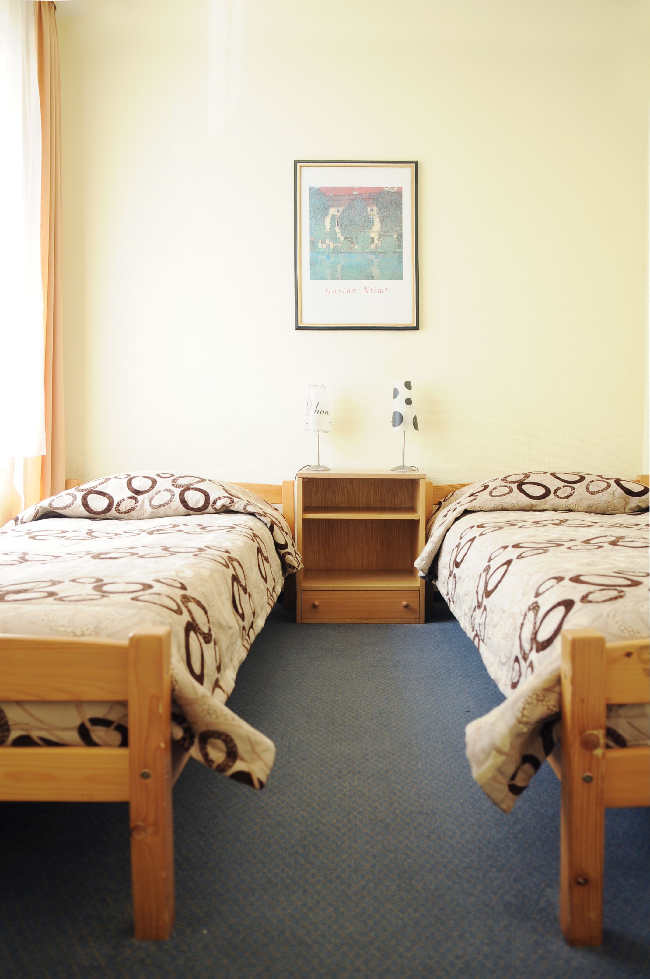 Een kamer met twee houten bedden, een klein houten nachtkastje met een lamp en een lijst met een foto aan de muur.