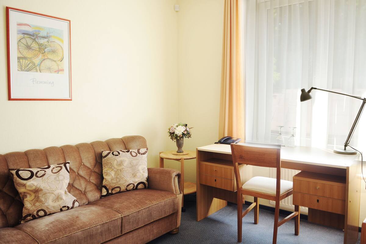 Ein Zimmer mit braunem Sofa, einem Holztisch, einem Stuhl, einem gerahmten Bild an der Wand und einem Fenster mit Vorhängen.
