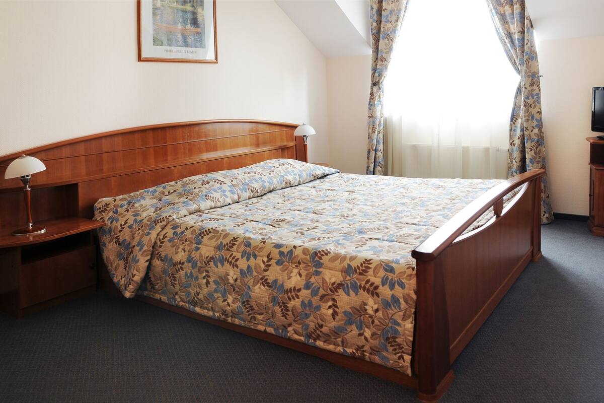 Ein Schlafzimmer mit einem hölzernen Bett, einem Nachttisch, einer Lampe, einem Fernseher und einem Fenster mit Vorhängen.