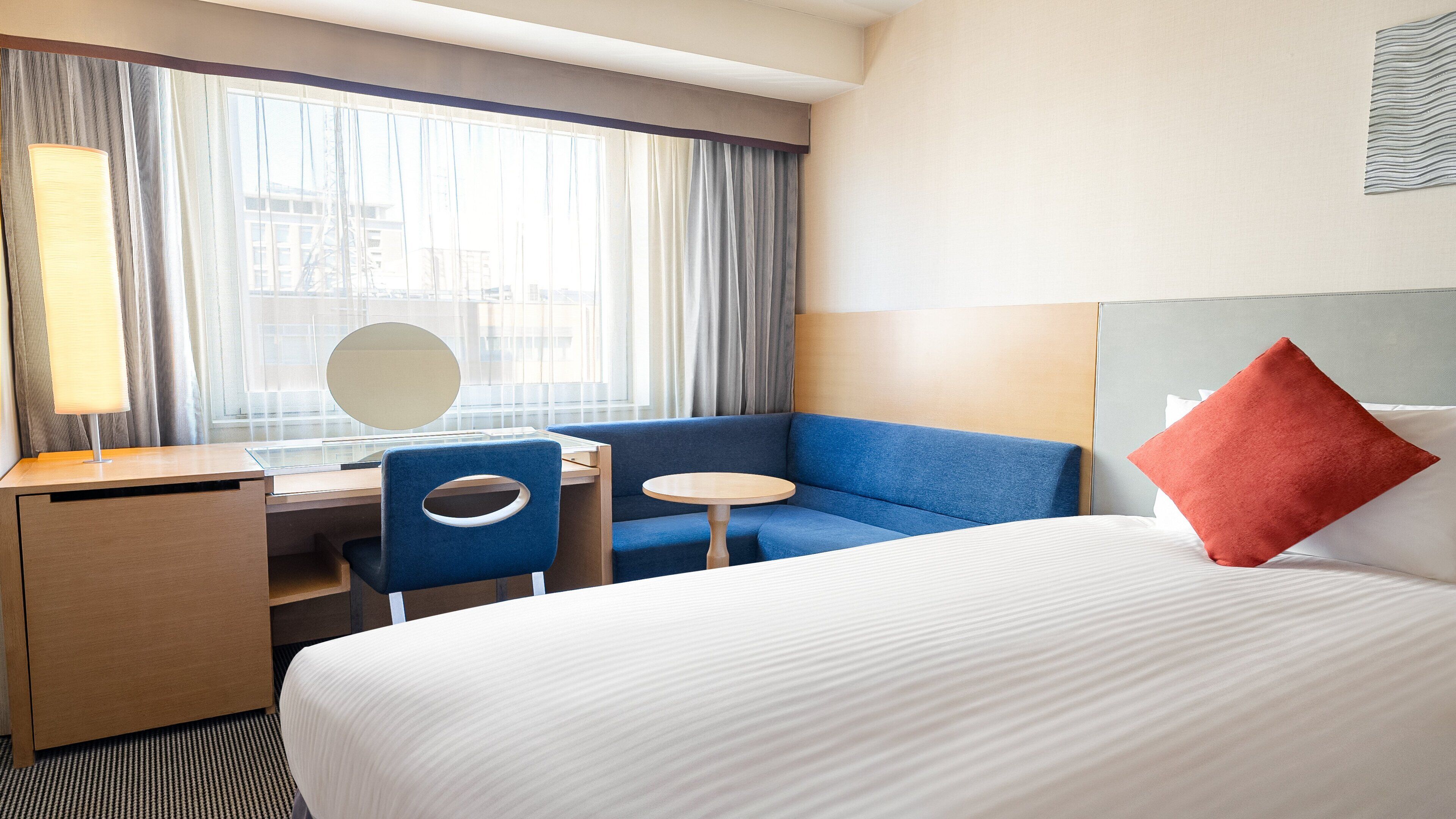 A hotel room with a bed, a desk, a chair, a small round table, and a large window with curtains.