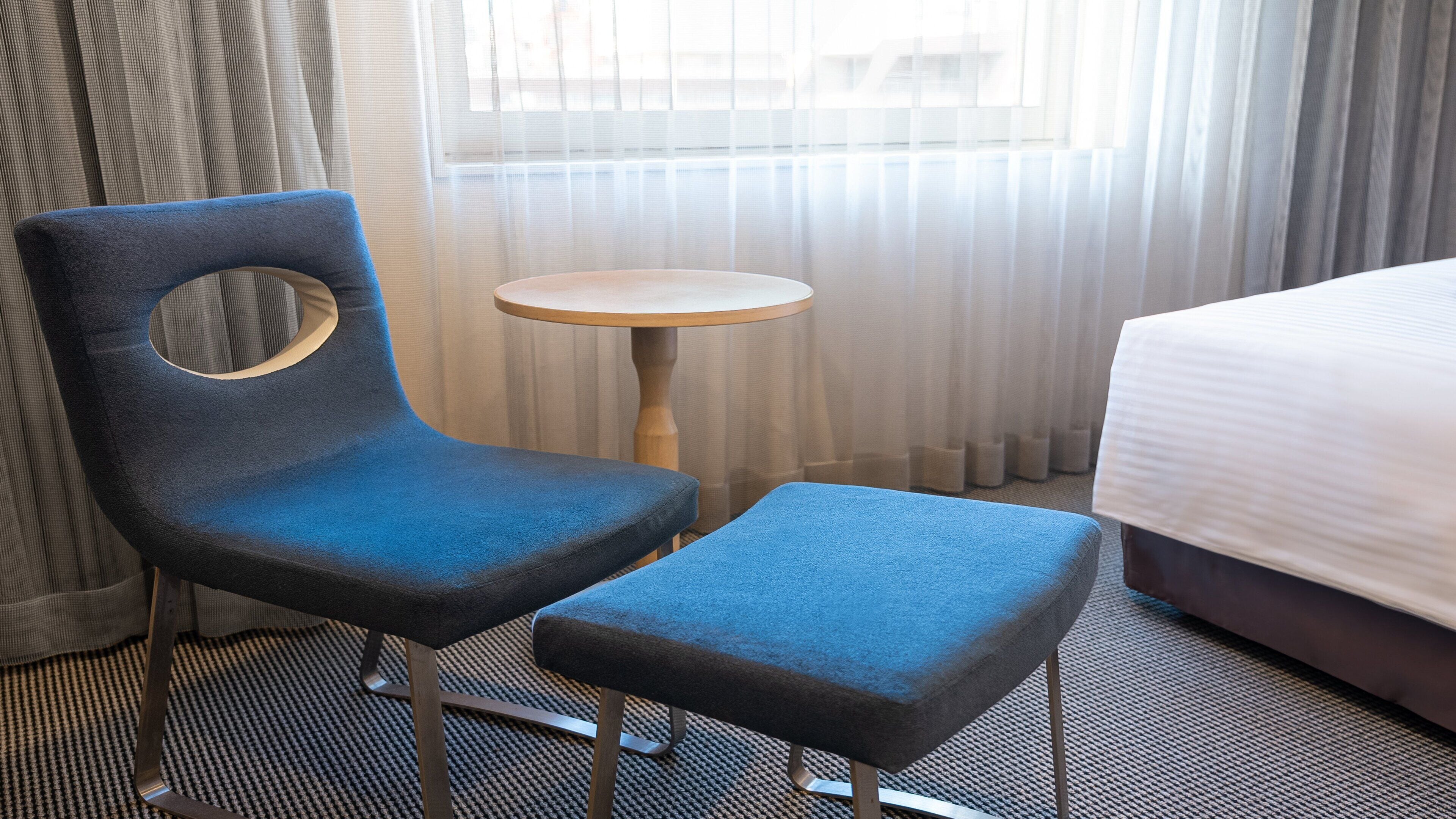 Een hotelkamer met een bed, een blauwe fauteuil met een ronde uitsparing, een klein rond tafeltje en dunne gordijnen.