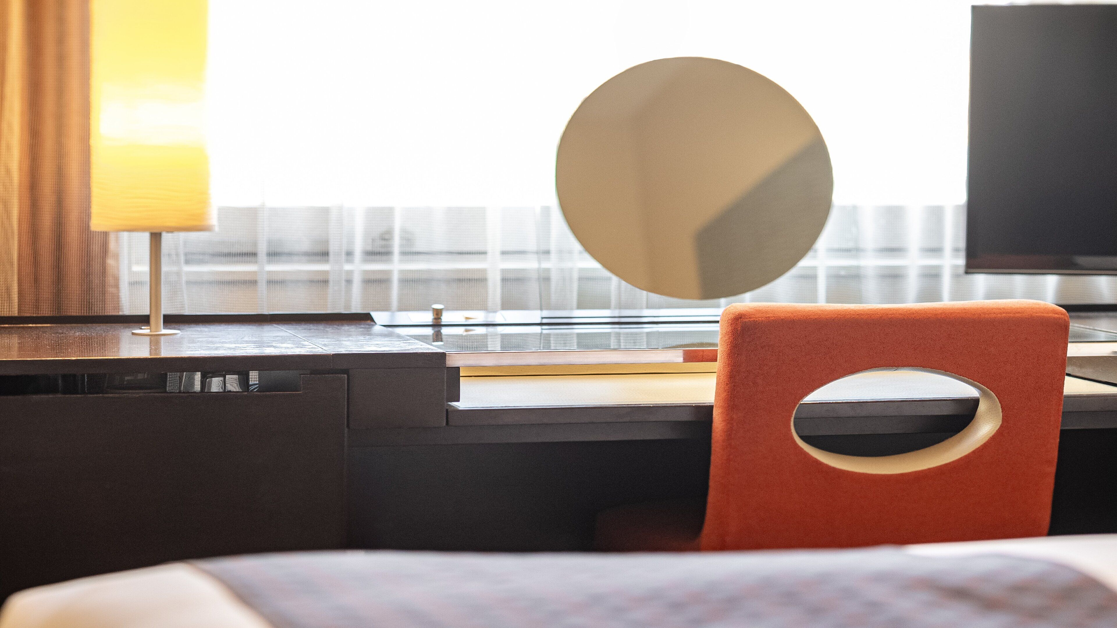 A modern hotel room with a desk, a chair, a large mirror, and a television.