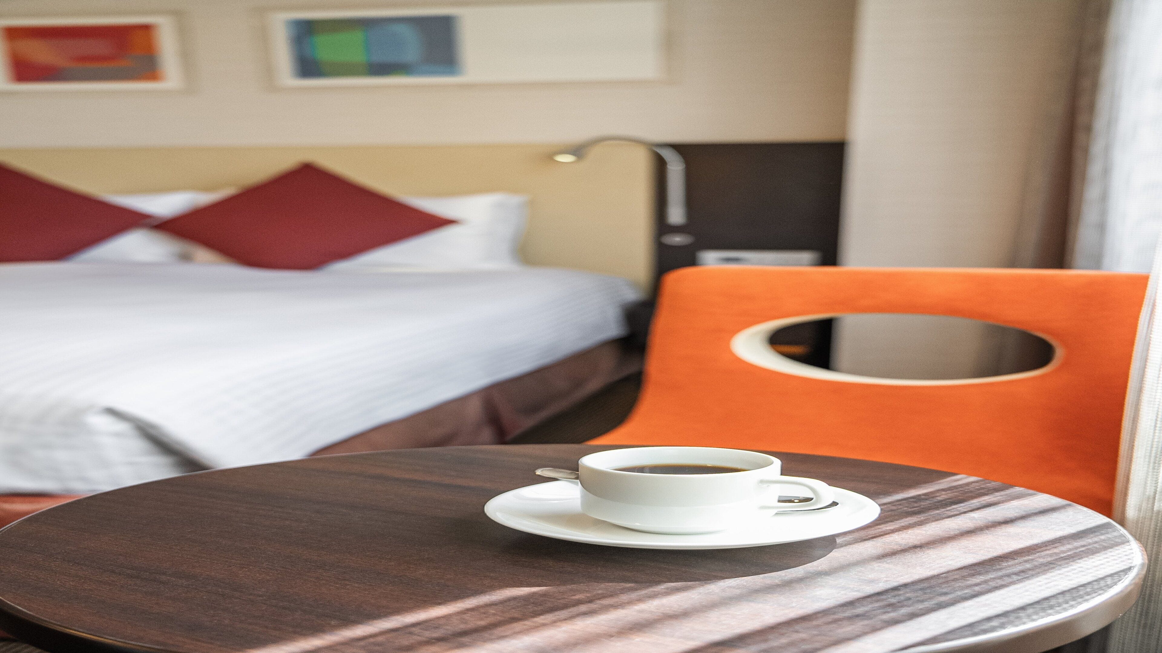 A hotel room with a bed, a table with a cup of coffee, and an orange chair.