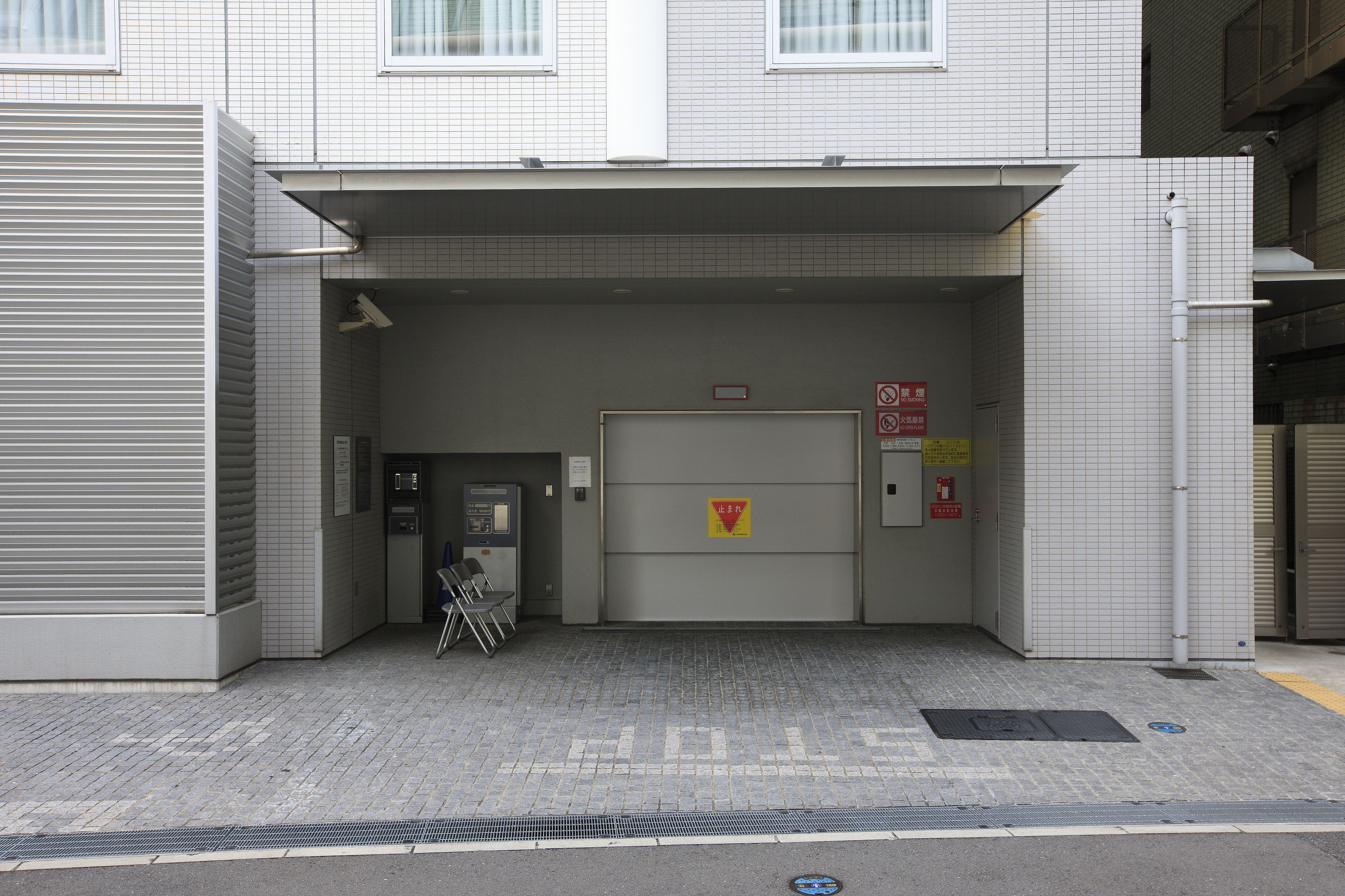 Ein Gebäudeeingang mit einer Kachelfassade, einer Tür mit einem roten und gelben Schild und einer kleinen Sitzecke mit Stühlen.