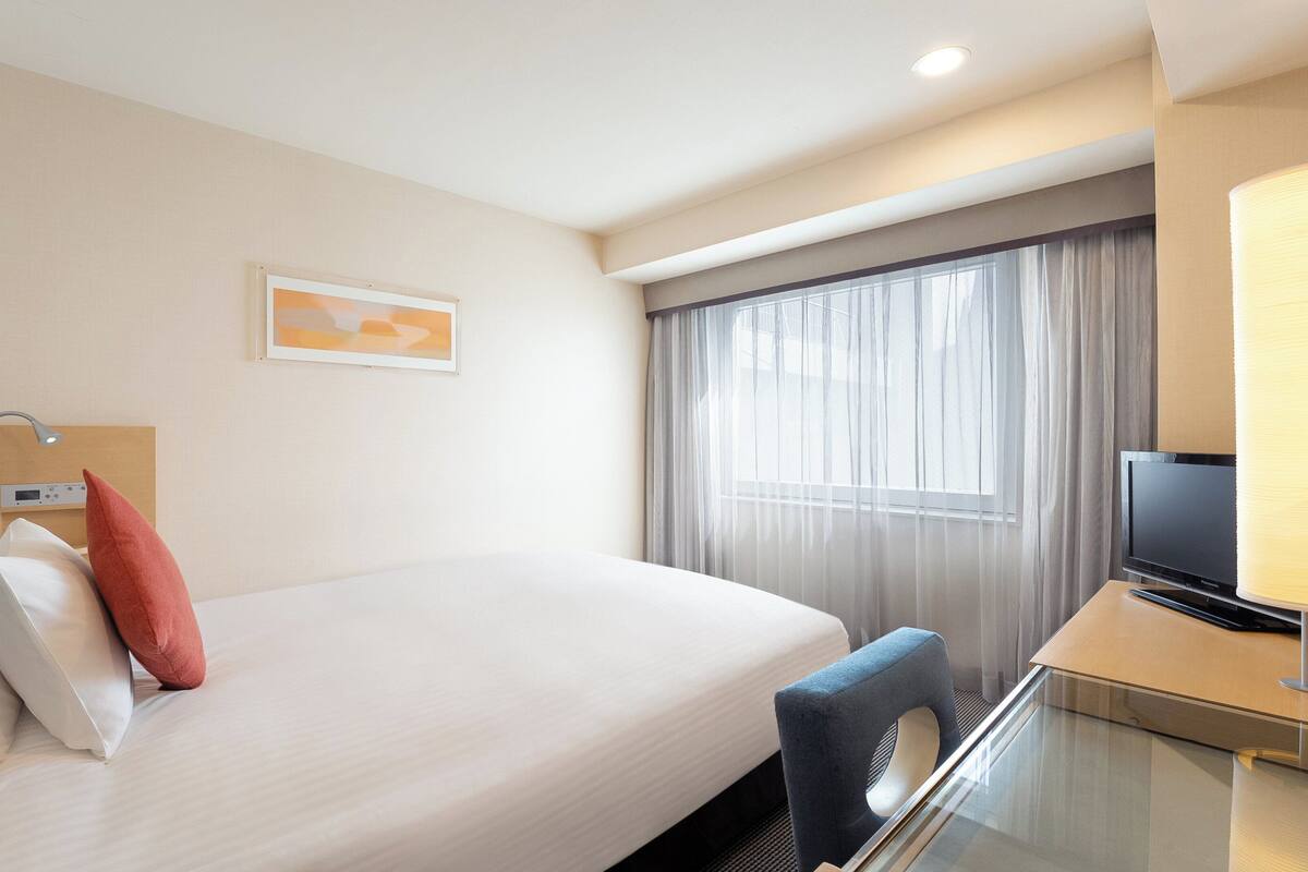 A hotel room with a bed, a desk with a television, a window with curtains, and a wall-mounted control panel.