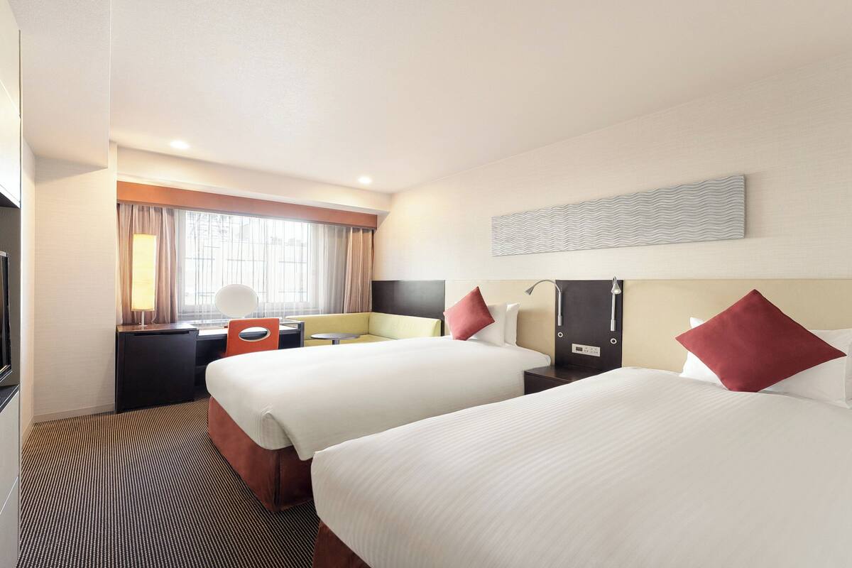 A hotel room with two beds, a desk, a chair, a TV, and a window with curtains.