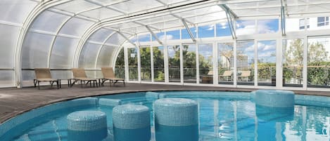 An indoor swimming pool with clear glass walls, lounge chairs, and a view of the outside.