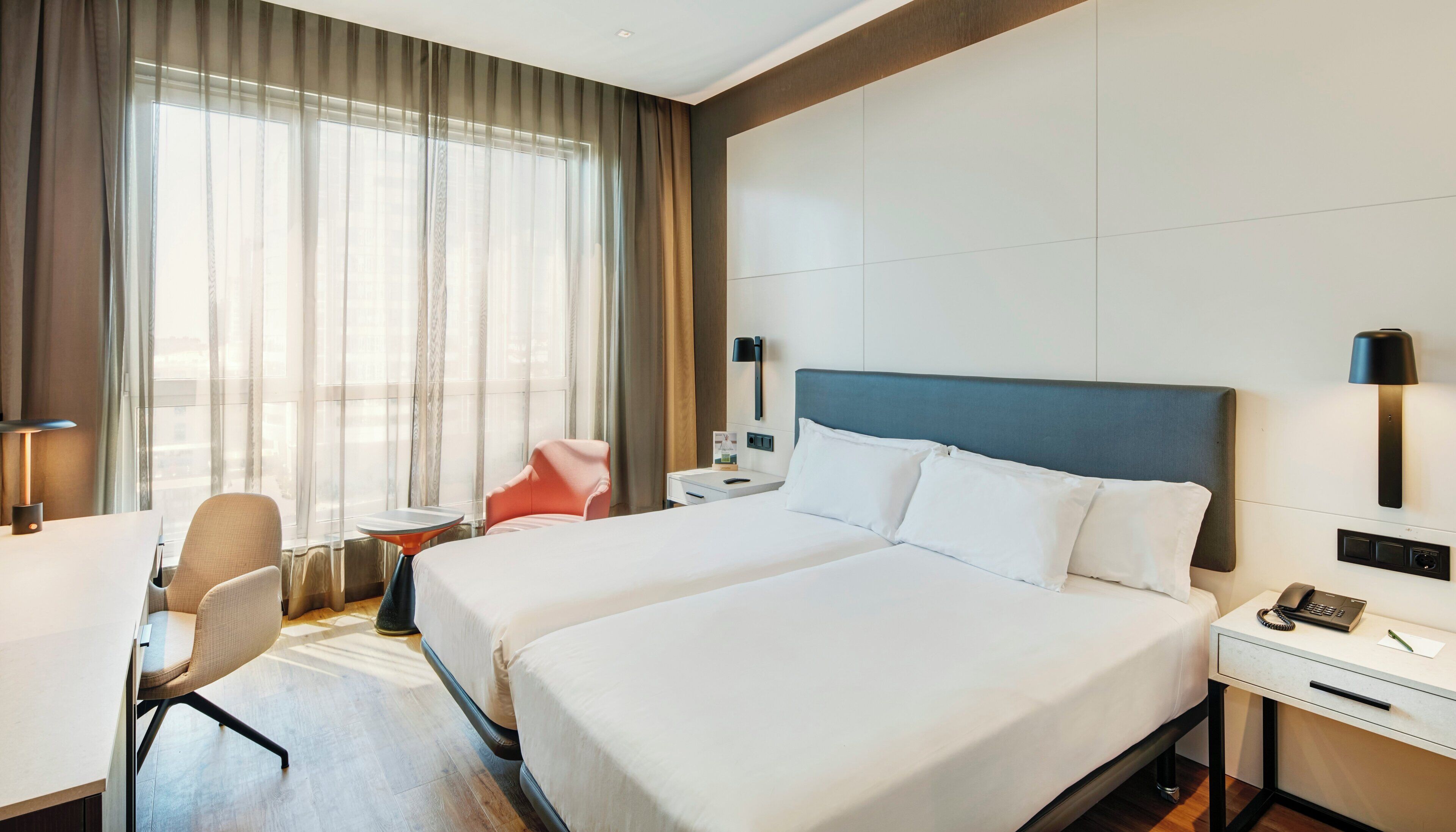 A hotel room with two beds, a desk, a chair, a small table, a lamp, and a nightstand with a phone.