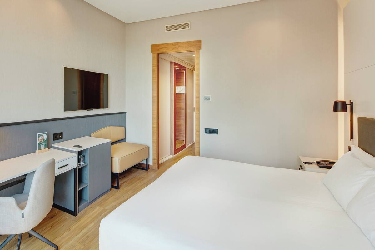 A hotel room with a bed, desk, chair, TV, and a door leading to another room.