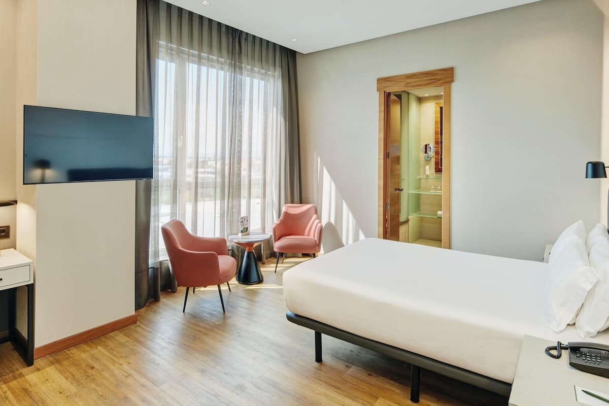 A hotel room with a bed, desk, chairs, a TV, and a bathroom door.