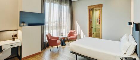 A hotel room with a bed, desk, chairs, a TV, and a bathroom door.