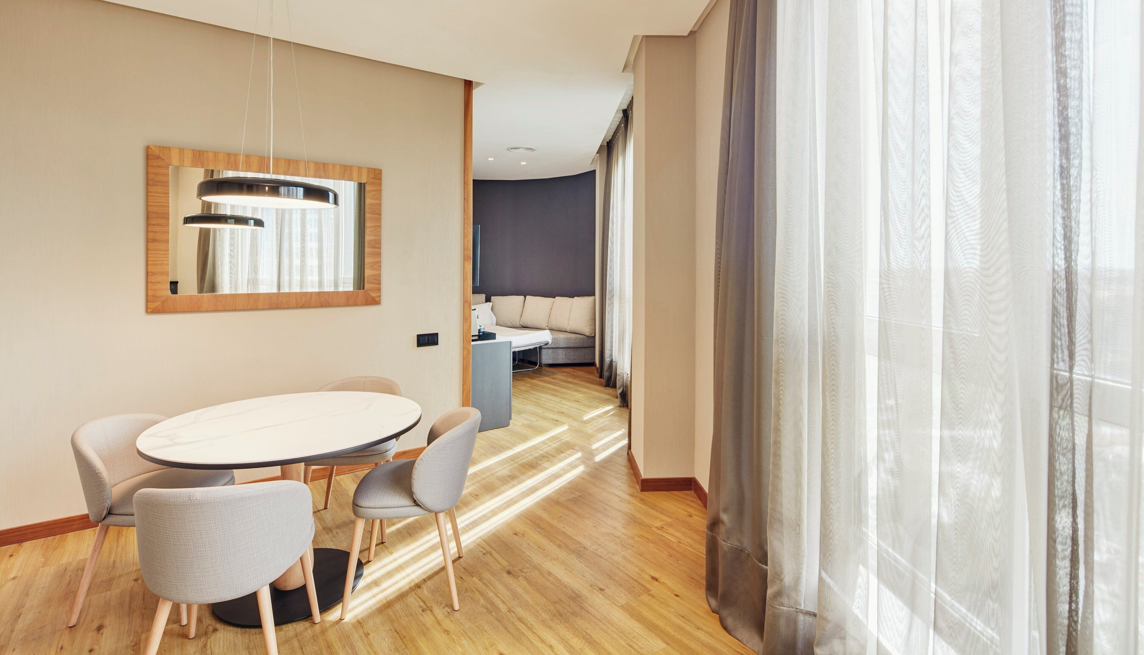 Ein modernes Hotelzimmer mit einem runden Esstisch, vier Stühlen, einer Couch, einem kleinen Schreibtisch und einem großen Fenster mit transparenten Vorhängen.