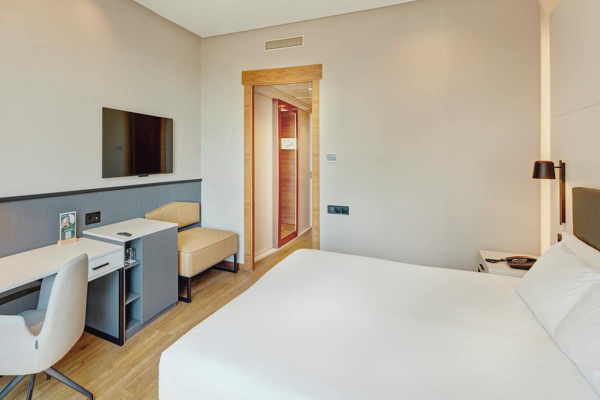 Une chambre d’hôtel équipée d’un lit, d’un bureau, d’une chaise, d’une télévision et d’une porte donnant sur une autre pièce.