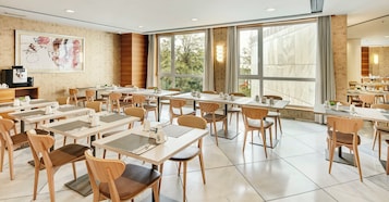 Un comedor moderno con varias mesas y sillas, ventanas amplias con vista a árboles y una máquina de café montada en la pared.