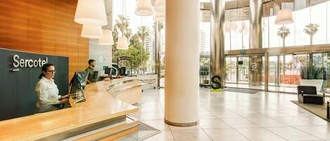 A hotel reception area with a modern design, featuring a reception desk, large columns, and a view of palm trees through the windows.