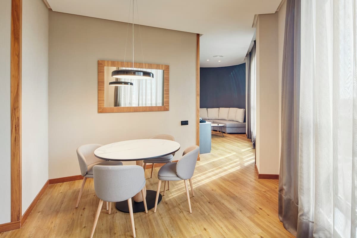 A modern hotel room with a dining area, a sofa, and a large window with curtains.