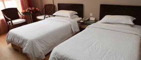 Two single beds with white linens, a wooden nightstand with a telephone, and a chair with a vase of flowers.