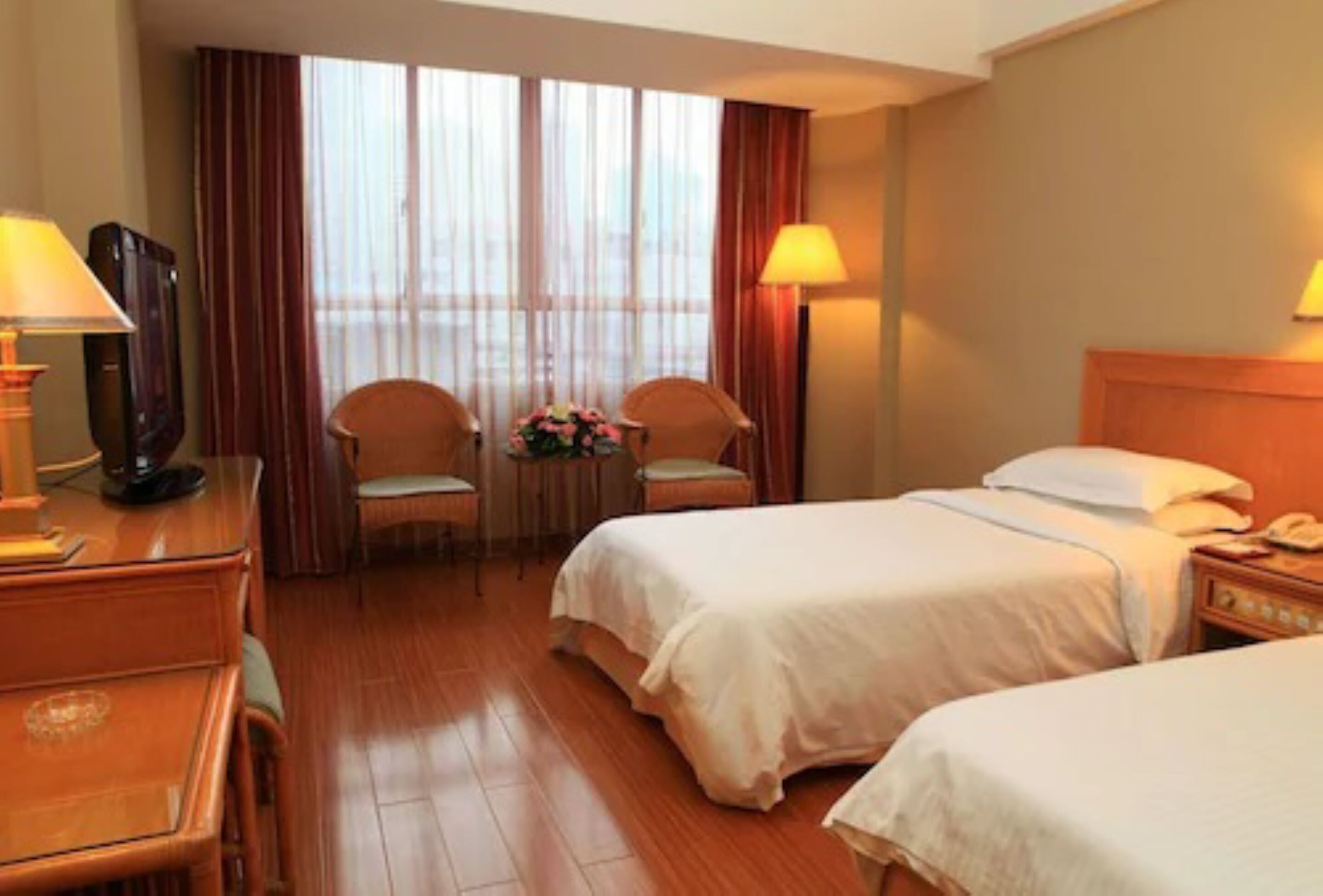 A hotel room with two beds, a desk with a lamp, a chair, and a small table with flowers.