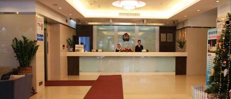 A hotel reception area with a glass counter, two receptionists, a red carpet, and a Christmas tree.