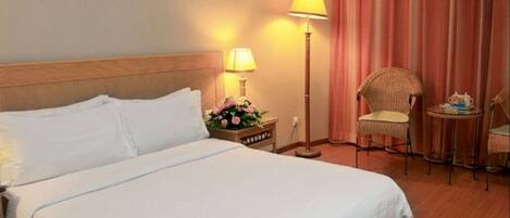 Quarto de hotel com uma cama grande, uma mesa de cabeceira com abajur e flores, uma cadeira e uma pequena mesa com um bule.