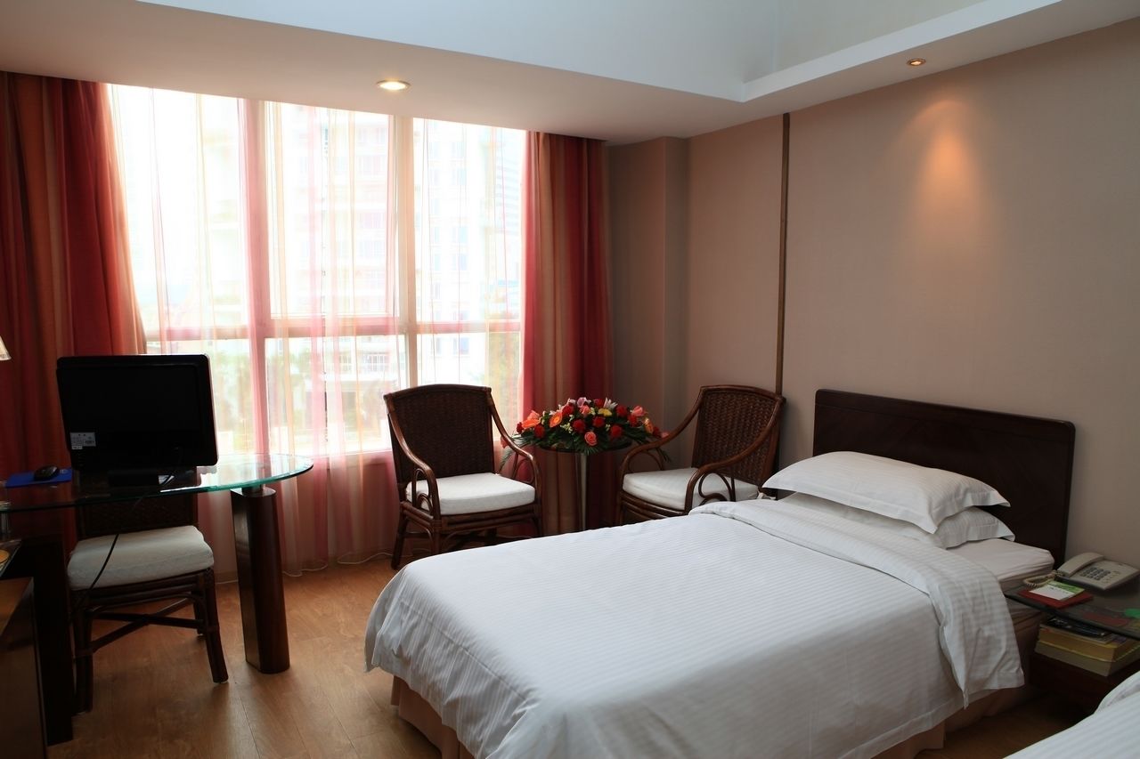A hotel room with a bed, a desk with a television, two chairs, a small table with flowers, and a bedside table with a phone and books.