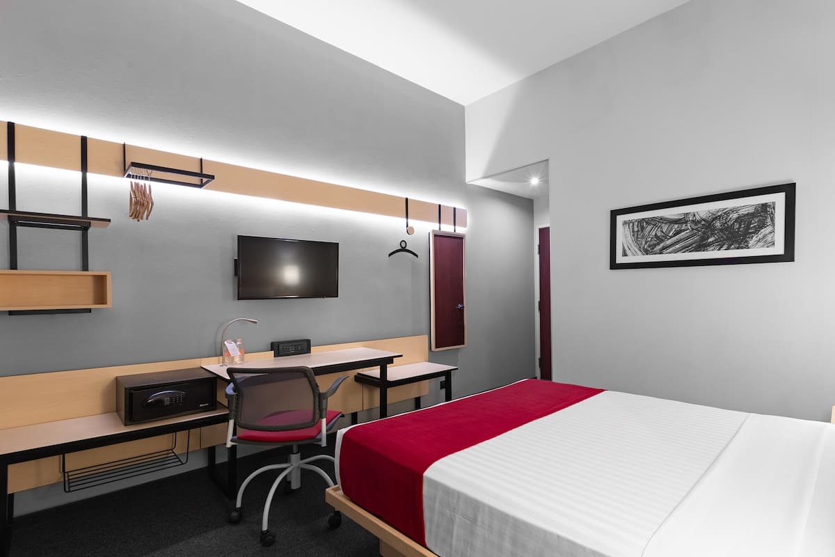 A modern hotel room with a bed, desk, chair, TV, and artwork on the wall.
