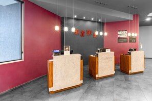 A reception area with three wooden reception desks, a red wall, and a grey wall with hanging lights. - City Express Plus by Marriott Guadalajara Expo (Zapopan)