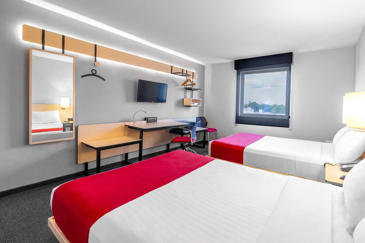 A hotel room with two beds, a desk, a TV, and a window.