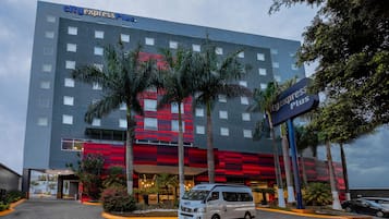 Un hotel City Express Plus con una fachada moderna, palmeras y una camioneta estacionada al frente.